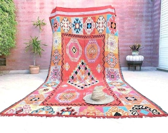 Puede incluir: Una alfombra rectangular grande con una base roja vibrante, un patrón central de diamantes y varias formas geométricas en naranja, rosa, azul y negro. Un sombrero de paja descansa sobre la superficie de la alfombra, que tiene flecos abajo.