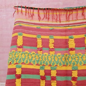 May include: A colorful woven throw blanket with a striped pattern in shades of pink, yellow, and green. The blanket has a textured surface with raised pom-poms.