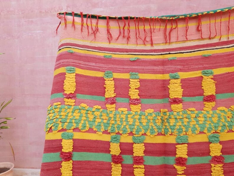 May include: A colorful woven throw blanket with a striped pattern in shades of pink, yellow, and green. The blanket has a textured surface with raised pom-poms.
