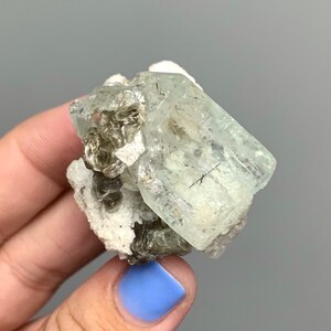 Puede incluir: Un cristal de aguamarina transparente con un tono verde claro, rodeado de un grupo de cristales grises m&aacute;s peque&ntilde;os en una matriz blanca.