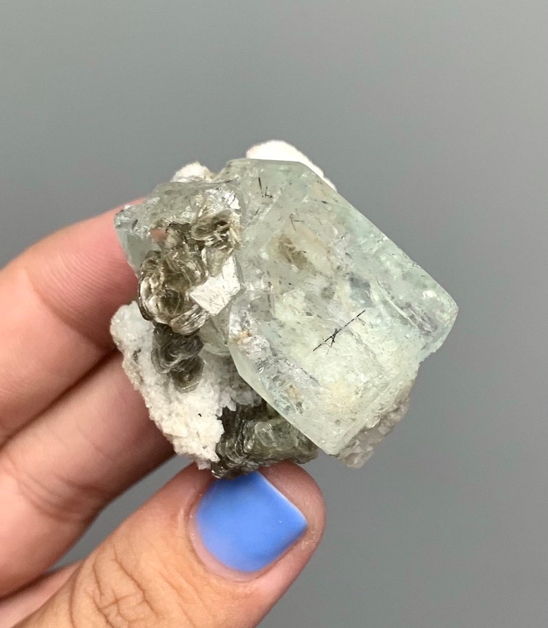 Puede incluir: Un cristal de aguamarina transparente con un tono verde claro, rodeado de un grupo de cristales grises m&aacute;s peque&ntilde;os en una matriz blanca.