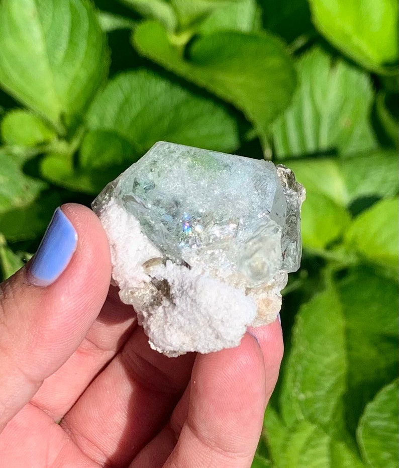 Puede incluir: Un cristal de aguamarina azul claro con una matriz blanca. El cristal est&aacute; facetado y tiene un ligero brillo iridiscente.