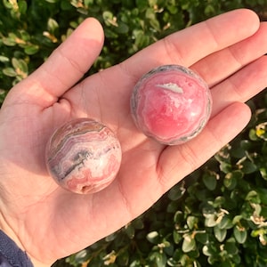 May include: Two pink and white striped spheres of polished pink calcite, also known as  pink manganese calcite, are shown in a hand. The spheres are different sizes.