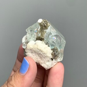 Puede incluir: Un grupo de cristales de fluorita verde claro con una matriz blanca. Los cristales de fluorita son transl&uacute;cidos y tienen un brillo v&iacute;treo. La matriz est&aacute; cubierta de peque&ntilde;os cristales grises.