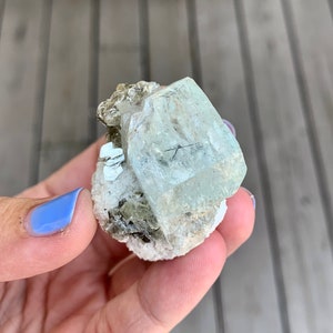Puede incluir: Un cristal de aguamarina verde claro con una textura clara y v&iacute;trea. El cristal est&aacute; rodeado de cristales marrones m&aacute;s peque&ntilde;os y est&aacute; colocado sobre una matriz blanca.