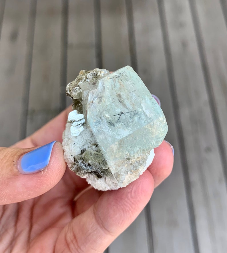 Puede incluir: Un cristal de aguamarina verde claro con una textura clara y v&iacute;trea. El cristal est&aacute; rodeado de cristales marrones m&aacute;s peque&ntilde;os y est&aacute; colocado sobre una matriz blanca.