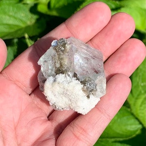Puede incluir: Un cristal transparente y facetado con una base blanca y rugosa. El cristal tiene un tono verde claro y est&aacute; rodeado de cristales grises m&aacute;s peque&ntilde;os.