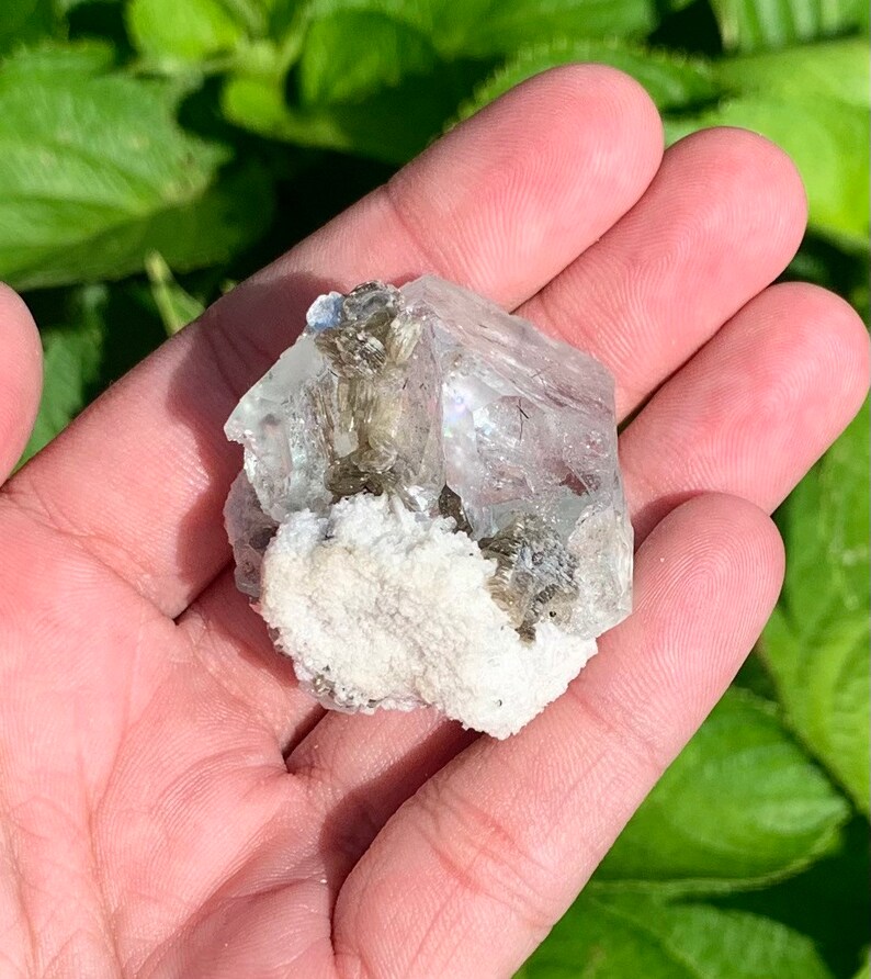 Puede incluir: Un cristal transparente y facetado con una base blanca y rugosa. El cristal tiene un tono verde claro y est&aacute; rodeado de cristales grises m&aacute;s peque&ntilde;os.