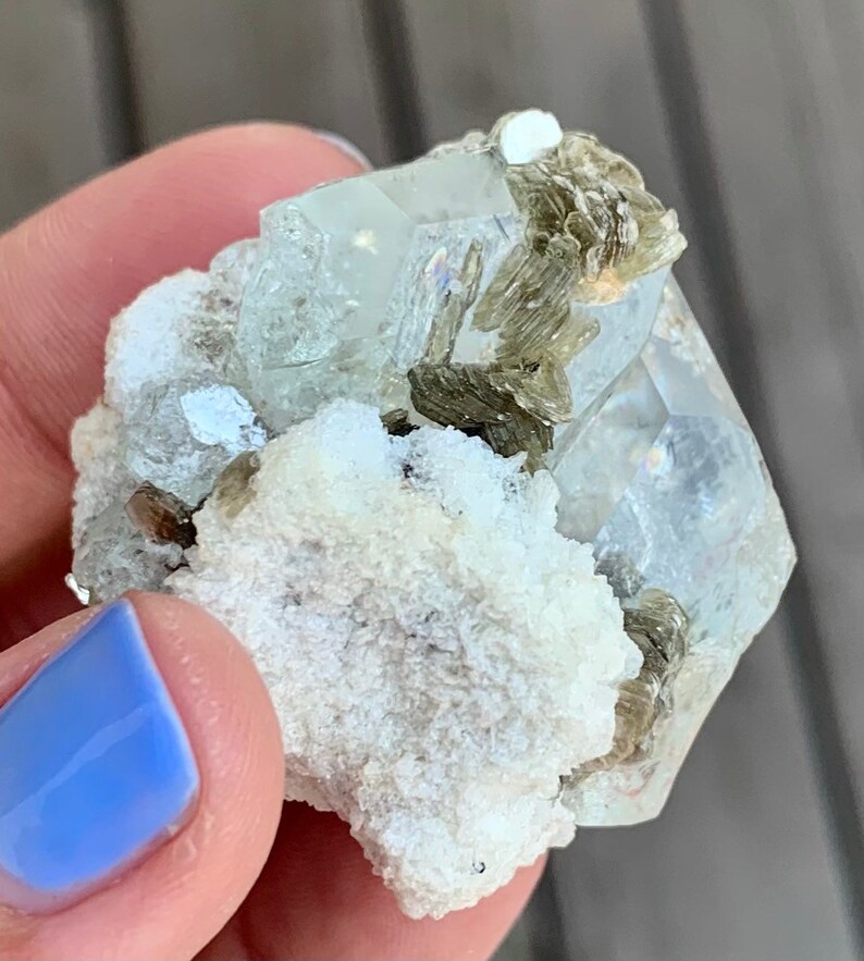 Puede incluir: Un c&uacute;mulo de cristales de aguamarina transparentes y facetados con inclusiones verdes, engastado en una matriz blanca. El cristal mide aproximadamente 5 cm.