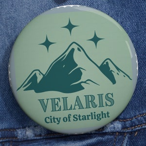 May include: A round, light green button with a mountain and star design. The text "VELARIS City of Starlight" is printed in dark green. The button is pinned to a blue denim jacket.