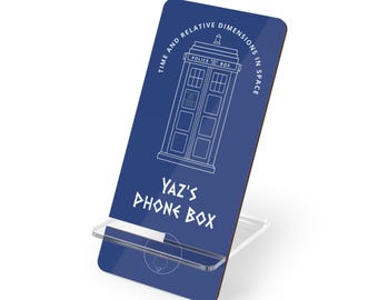 Puede incluir: Un soporte para teléfono azul con un contorno blanco de una cabina de policía y el texto "TIME AND RELATIVE DIMENSIONS IN SPACE" y "YAZ'S PHONE BOX". El soporte tiene una pieza rectangular azul y una base de plástico transparente.