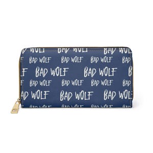 May include: A navy blue wallet with a gold zipper closure. The wallet is covered in a repeating pattern of the words "BAD WOLF" in white, handwritten-style lettering. The wallet is rectangular and appears to be made of a leather-like material.
