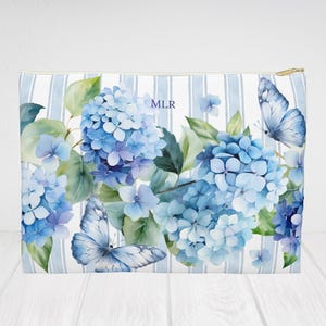 Puede incluir: Un neceser rectangular con un fondo a rayas azules y blancas. La bolsa presenta un diseño de acuarela de hortensias azules y mariposas. Las iniciales "MLR" están impresas en la bolsa.