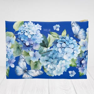 Op de afbeelding: Een blauwe etui met ritssluiting met een aquarelontwerp van blauwe hortensia's en vlinders. De etui heeft een gouden rits en de initialen "MLR" zijn zichtbaar. De achtergrond is helderblauw.