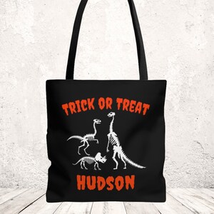 Puede incluir: Bolso tote negro con gráficos de esqueletos de dinosaurios blancos y el texto "TRICK OR TREAT" en naranja. Debajo está el nombre "HUDSON" en naranja. El bolso tiene asas negras.