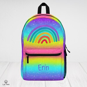 Puede incluir: Una mochila con un fondo de degradado de arcoíris vibrante y un patrón de estrellas. El frente muestra un gráfico de arcoíris colorido y el nombre Erin impreso en texto morado. Incluye un asa superior negra, cremalleras negras y un bolsillo frontal.