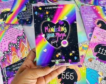 May include: A 52-card affirmation deck in a clear plastic case with a black heart-shaped center featuring the text "Manifesting Your Magic" and "by Timmery" surrounded by a vibrant rainbow gradient, small colorful hearts, and stars.