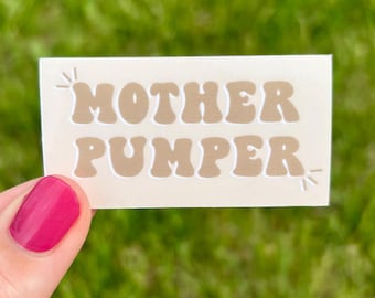 May include: A rectangular white sticker featuring the text MOTHER PUMPER in a wavy, bold, tan-colored font. The design includes small, simple starburst accents in the top left and bottom right corners of the sticker, which has a smooth finish.