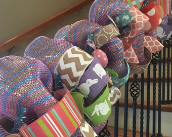 May include: A pink, purple, green, brown, and white Easter wreath with bunny and egg designs. The wreath is made of mesh and ribbon and is decorated with various Easter-themed embellishments.