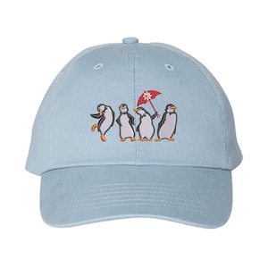 May include: A light blue baseball cap featuring an embroidered graphic of four penguins standing in a row. One penguin holds a small red umbrella with a white flower design on top. The cap has a curved brim and a structured six-panel crown design.