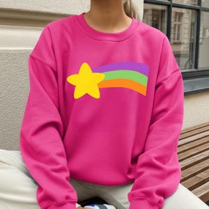 May include: A bright pink sweatshirt featuring a yellow star and a rainbow-colored stripe design. The star is on the left side, with the rainbow stripes extending to the right. The sweatshirt is a solid color, with a crew neck.