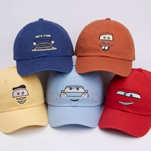May include: A collection of five baseball caps in blue, orange, yellow, light blue, and red. Each cap features an embroidered cartoon car face on the front panel, including eyes, grilles, and bumpers, with a curved brim and a structured crown.
