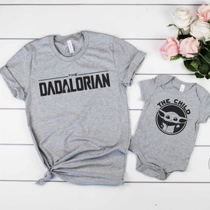 May include: Two grey t-shirts with a Star Wars theme. The larger shirt says "The Dadalorian" in black text. The smaller shirt says "The Child" in black text and features a black and white image of a character from the show.