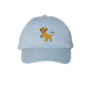 May include: A light blue baseball cap featuring an embroidered graphic of a young lion character with golden fur, a pink nose, and a long tail. The cap has a structured crown, a curved brim, and a button at the top center of the fabric panels.