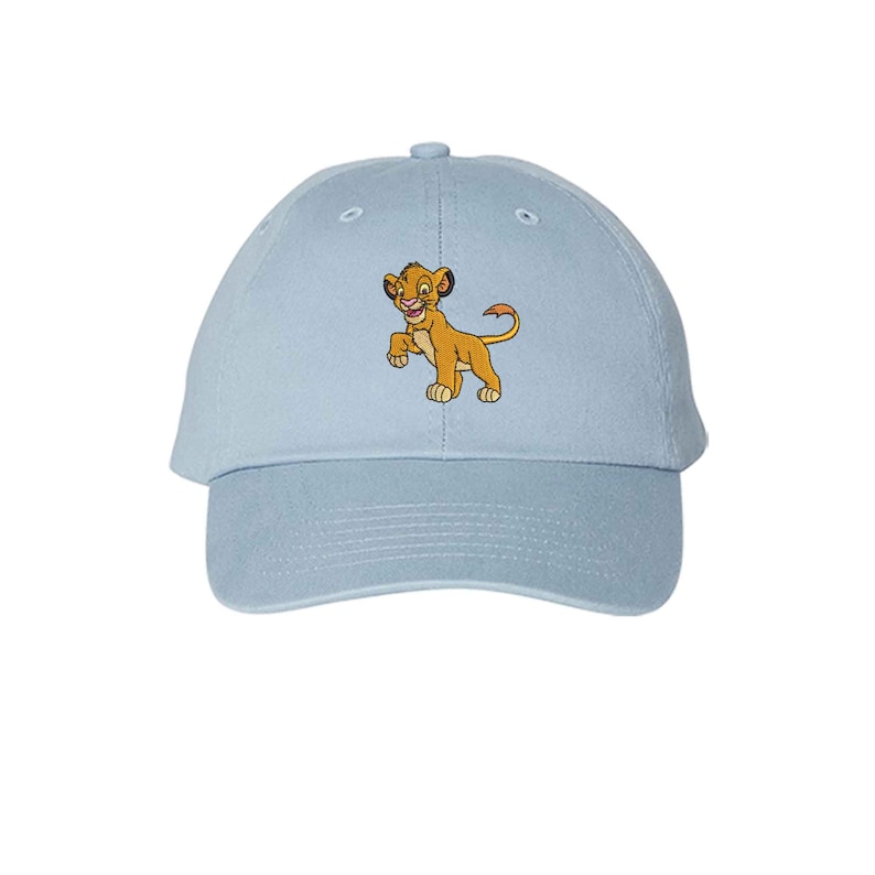 May include: A light blue baseball cap featuring an embroidered graphic of a young lion character with golden fur, a pink nose, and a long tail. The cap has a structured crown, a curved brim, and a button at the top center of the fabric panels.
