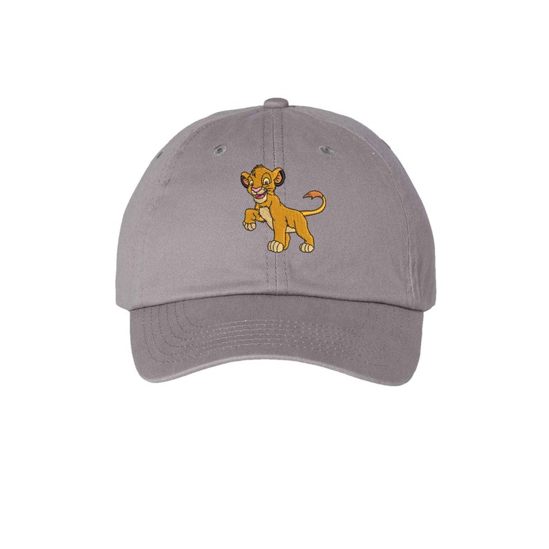 May include: A grey baseball cap featuring an embroidered graphic of a small, golden-brown lion cub standing on its hind legs. The cap has a curved brim, a button on top, and stitched eyelets for ventilation, providing a classic structured look.