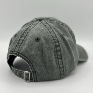 May include: A charcoal gray baseball cap with a washed, distressed fabric finish. The cap features a six-panel construction with stitched eyelets and a metal sliding buckle closure on the back strap for an adjustable fit and a curved brim.