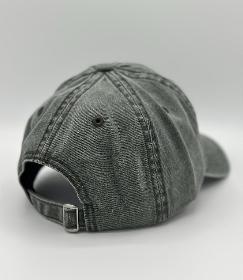 May include: A charcoal gray baseball cap with a washed, distressed fabric finish. The cap features a six-panel construction with stitched eyelets and a metal sliding buckle closure on the back strap for an adjustable fit and a curved brim.