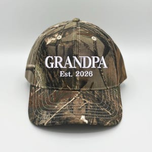 May include: A camouflage baseball cap featuring a woodland pattern in shades of brown, green, and black. The front of the cap is embroidered with the white text "GRANDPA" in a bold font, followed by the white text "Est. 2026" underneath.
