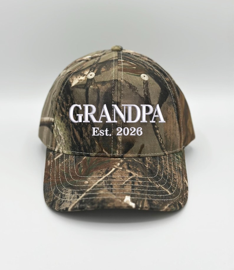 May include: A camouflage baseball cap featuring a woodland pattern in shades of brown, green, and black. The front of the cap is embroidered with the white text "GRANDPA" in a bold font, followed by the white text "Est. 2026" underneath.