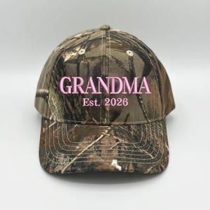 May include: A camouflage baseball cap featuring the embroidered text GRANDMA Est. 2026 in light pink thread. The cap has a structured crown, a curved brim with visible stitching, and a woodland-inspired pattern in shades of brown, green, and tan.