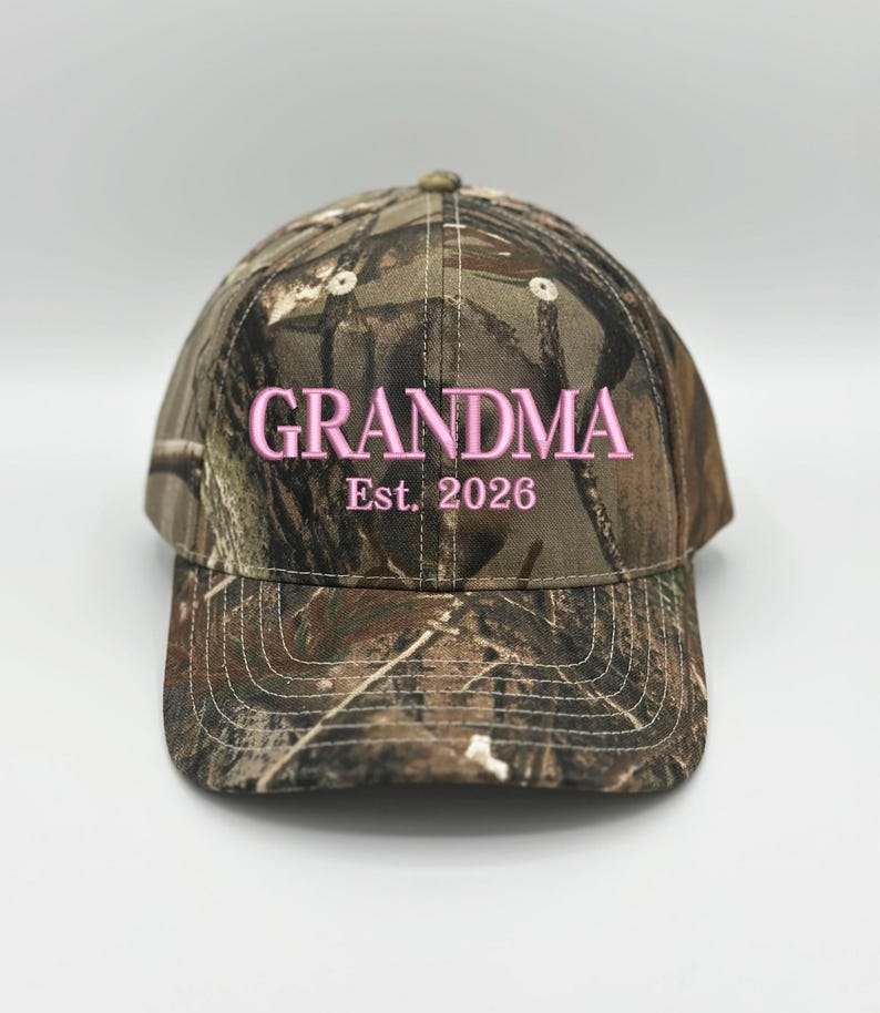 May include: A camouflage baseball cap featuring the embroidered text GRANDMA Est. 2026 in light pink thread. The cap has a structured crown, a curved brim with visible stitching, and a woodland-inspired pattern in shades of brown, green, and tan.