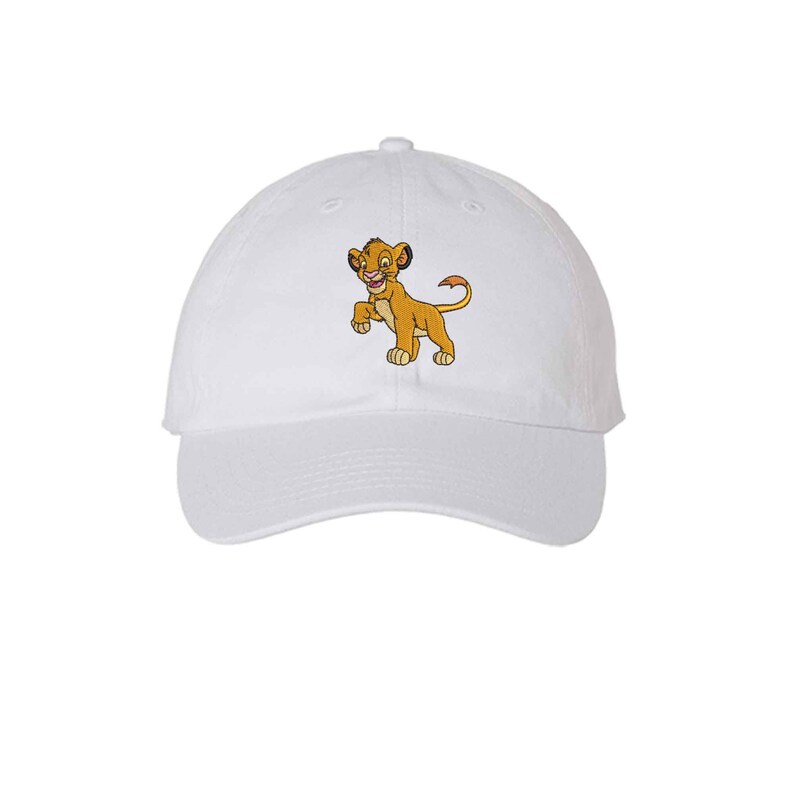 May include: A white baseball cap featuring an embroidered graphic of a young lion character with golden-brown fur, a pink nose, and a tufted tail. The cap has a curved brim and a structured crown, showcasing a detailed depiction of the cub.