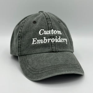 May include: A charcoal gray baseball cap with a washed, distressed fabric finish. The front panel features the words Custom Embroidery stitched in white thread. The cap has a curved brim, stitched eyelets, and a structured six-panel crown design.