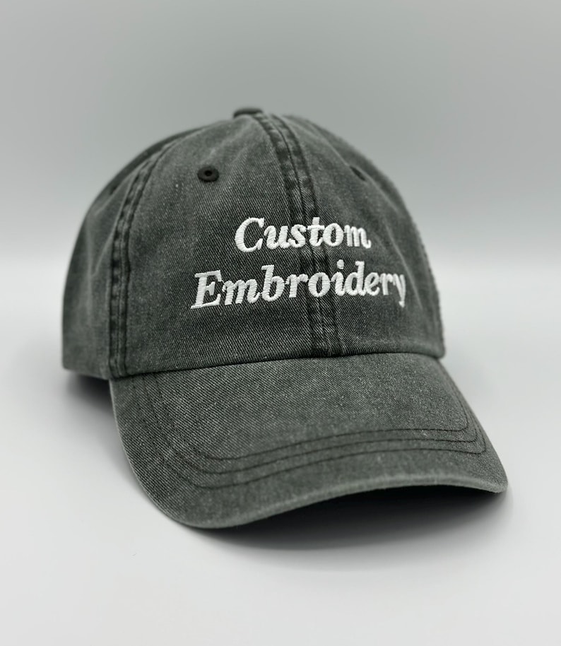 May include: A charcoal gray baseball cap with a washed, distressed fabric finish. The front panel features the words Custom Embroidery stitched in white thread. The cap has a curved brim, stitched eyelets, and a structured six-panel crown design.