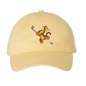 May include: A light yellow baseball cap featuring a cartoon tiger playing golf. The tiger is orange and black, holding a golf club, with a golf ball and green grass detail. The cap has a curved brim and a button on top.
