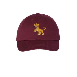 May include: A maroon baseball cap featuring an embroidered graphic of a young lion cub with golden fur, a light-colored belly, and a tufted tail. The cap has a curved brim, a button on the top center, and stitched eyelets for ventilation.