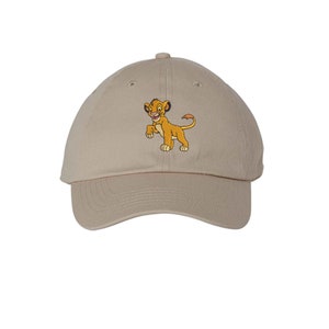 May include: A beige baseball cap featuring an embroidered graphic of a young lion cub. The cub is depicted in shades of yellow and orange with a pink nose, standing on all fours. The cap has a curved brim and a structured crown with eyelets.