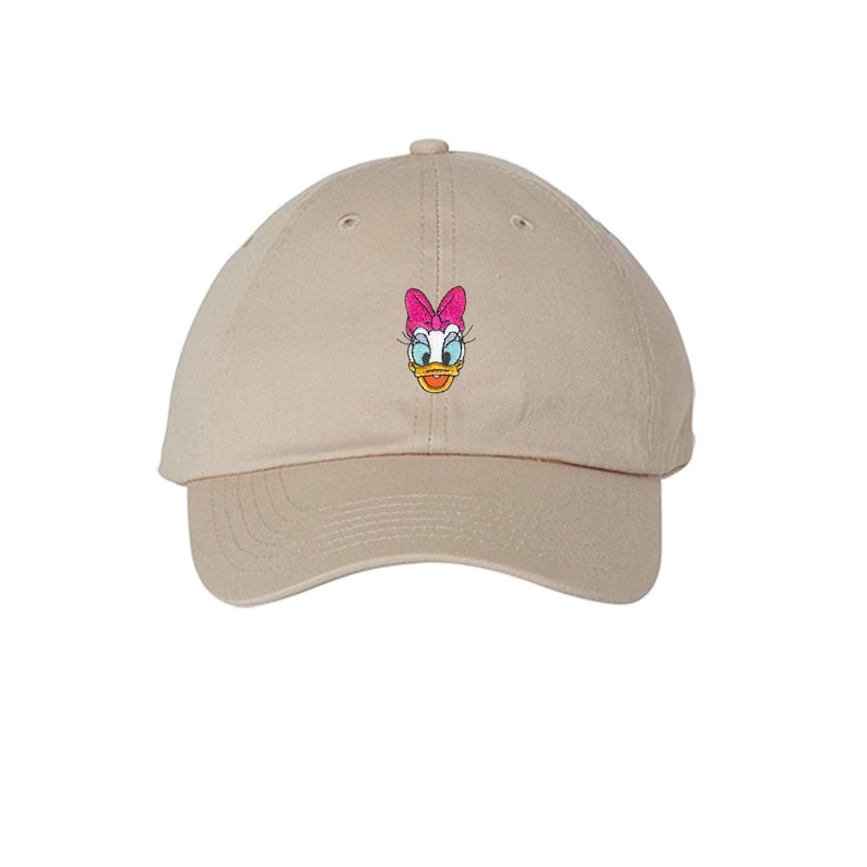 Op de afbeelding: Een beige baseballpet met een geborduurd beeld van Daisy Duck met een roze strik.