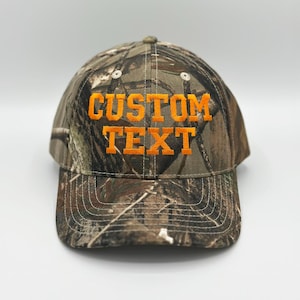 May include: A baseball cap featuring a camouflage pattern in shades of brown, green, and black. The front of the cap displays the words CUSTOM TEXT embroidered in bright orange block lettering. The cap has a curved brim and stitched eyelets.