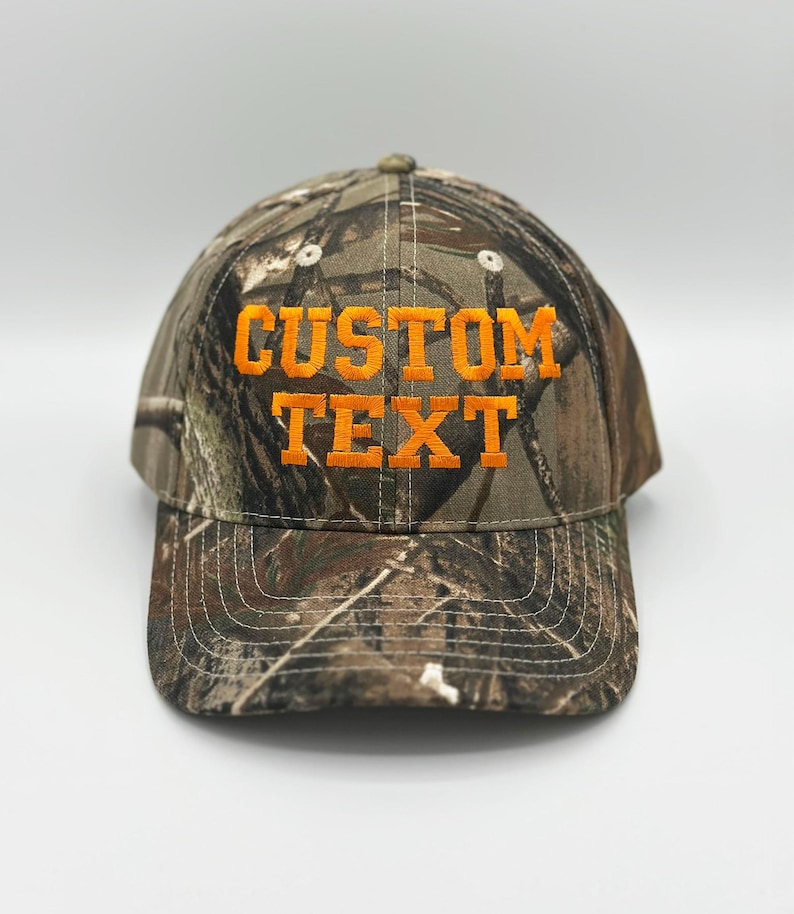 May include: A baseball cap featuring a camouflage pattern in shades of brown, green, and black. The front of the cap displays the words CUSTOM TEXT embroidered in bright orange block lettering. The cap has a curved brim and stitched eyelets.
