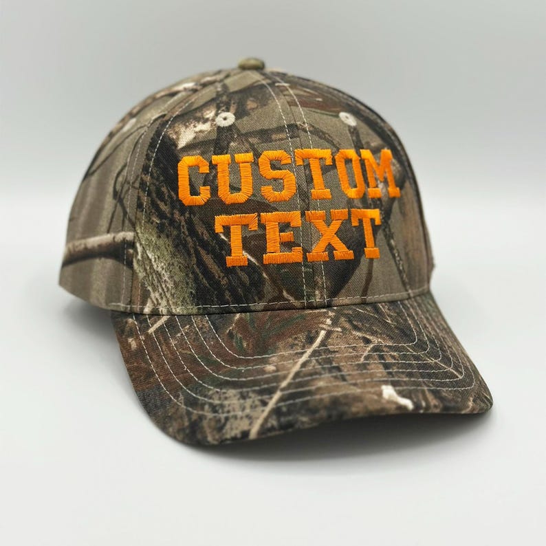 May include: A baseball cap featuring a camouflage pattern in shades of brown, green, and black. The front of the cap displays the words CUSTOM TEXT embroidered in bold, bright orange capital letters with a structured crown and a curved brim.