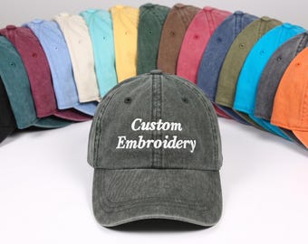 Op de afbeelding: Een antracietgrijze honkbalpet met de wit geborduurde tekst Custom Embroidery op het voorpaneel. De pet is geplaatst voor een halve cirkel van meerdere honkbalpetten in verschillende kleuren, waaronder blauw, rood, geel en groen.