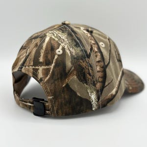 May include: A baseball cap featuring a brown, tan, and dark green camouflage pattern. The cap has a structured crown with stitched eyelets and a black plastic adjustable strap closure at the back. The fabric displays a detailed forest-inspired print.