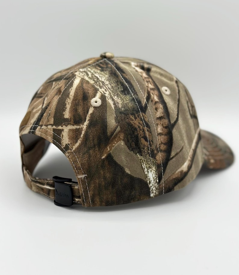 May include: A baseball cap featuring a brown, tan, and dark green camouflage pattern. The cap has a structured crown with stitched eyelets and a black plastic adjustable strap closure at the back. The fabric displays a detailed forest-inspired print.