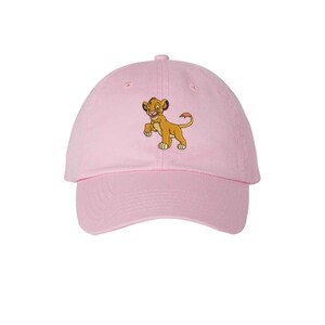May include: A light pink baseball cap featuring an embroidered graphic of a young lion cub with golden-brown fur, dark ear tips, and a tufted tail. The cap has a curved brim, a button on the top, and stitched eyelets for ventilation.